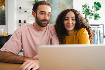 A young couple waving at their laptop screen, engaged in a friendly video call, with a colorful background, relaxed vibe, and potted plants around them in a comfortable room.