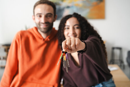 Close-up image of a couple’s hands holding a set of keys, with both smiling in the background, capturing the excitement of a new home or shared living space in a warmly lit room.
