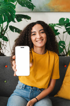 A smiling woman on a sofa, holding up a smartphone with the screen facing forward, in a comfortable, plant-filled room that exudes warmth and friendliness, creating a cozy atmosphere.