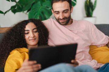 A couple comfortably leaning on a couch while browsing a tablet together, surrounded by a cozy, well-decorated living room with indoor plants, capturing a moment of shared relaxation.
