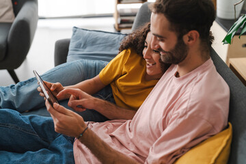 A couple comfortably leaning on a couch while browsing a tablet together, surrounded by a cozy, well-decorated living room with indoor plants, capturing a moment of shared relaxation.