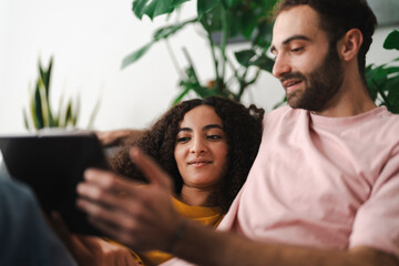 A couple sitting together on a couch, looking at a tablet in a bright and modern living room filled with greenery, enjoying a cozy and relaxed moment at home.