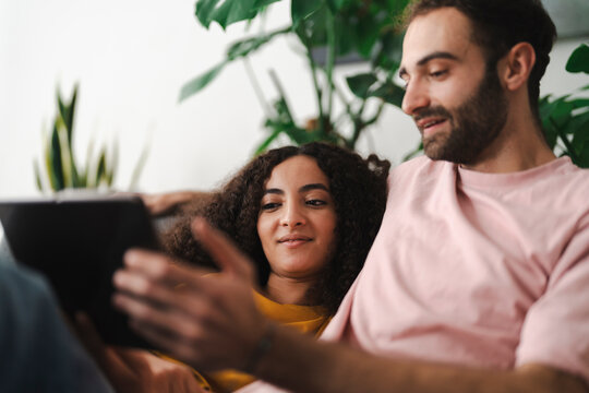 A couple sitting together on a couch, looking at a tablet in a bright and modern living room filled with greenery, enjoying a cozy and relaxed moment at home.