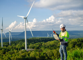 Young engineer looks at and inspects wind turbines on a plateau using a digital tablet.