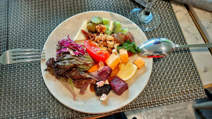 Top View Mixed Mediterranean Salad: Nutritious Flat Lay of Cheese, Purple Cabbage, Red Spinach, Tomatoes, Lemon, Kiwi, Olives, Dates, Hazelnuts, Cashews, Red Meat, Green Lettuce on Elegant White Plate