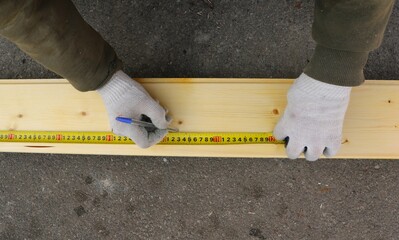 A man in work clothes and gloves uses a tape measure and pen to make markings on a light-colored wooden board lying on the asphalt