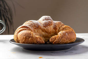 A freshly baked, golden-brown croissant with a dusting of powdered sugar rests on a sleek black plate, set on a white marble background with scattered crumbs.