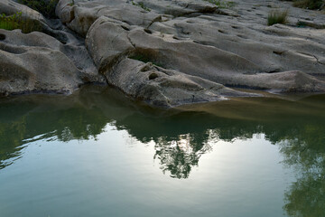 small pond in the middle of a rocky area, surrounded by lush green grass and plants. The water is still and clear, reflecting the trees and sky above.