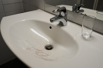 A white sink below a mirror in a bathroom. The sink has hair, a faucet, and a glass, and there are tiles on the wall behind it.