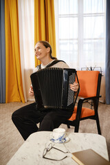 A middle-aged woman plays the accordion joyfully in a cozy room with colorful curtains and a warm ambiance