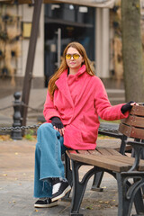 Woman in a pink coat and yellow sunglasses sitting on a bench in an urban park during fall