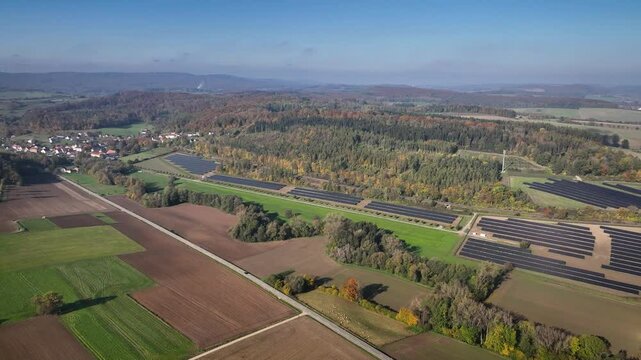 Aerial shot of solar park's structured layout with panels in varied symmetrical patterns among farmland and dense forest. Ground-mounted solar photovoltaic PV installations for renewable energy.