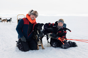 Couple enjoying mushing experience with sled dogs in the snowy arctic wilderness , on a winter adventure, dressed in warm gear, smiling, bonding with huskies, Tarvantovaara Wilderness Area, Finland