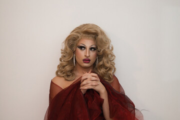 A drag queen with blonde wig and luxurious earrings and covered in red silk and on a white background with clasped hands praying imitating the virgin. Concept of lgtbqi rights and equality.