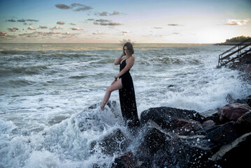 A beautiful young girl in a long black dress stands on large stones on the shore of a raging ocean.