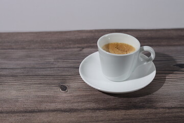 A white cup of espresso with its saucer on a wooden pattern with a white background.
