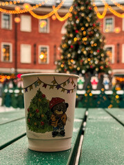 Cup with a bear on it sits on a table next to a Christmas tree. The cup is decorated with Christmas decorations and the bear is wearing a red hat. The scene is festive and cozy, with the cup