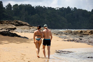 Couple in swimwear walking by the sand of tropical sea beach, back view. Holidays on sunny coast