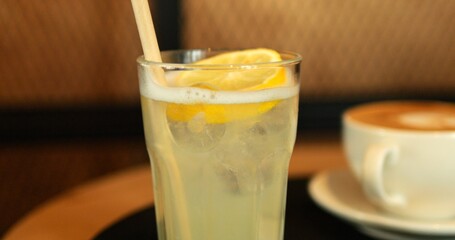 Fresh lemonade with straw and lemon slice near coffee cup on cafe table. Organic refreshing drink with citrus ingredients in glass. Exotic cocktail