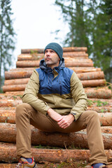Obraz premium middle-aged caucasian man in hat and jacket is sitting on the logs in the forest