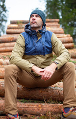 middle-aged caucasian man in hat and jacket is sitting on the logs in the forest