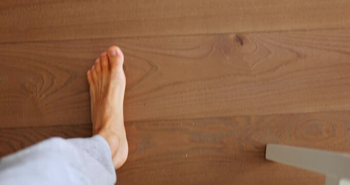 Man with bare feet walks on wooden floor of room with bright sun glare. Young guy in light grey trousers walks on parquet passing by chair slow motion