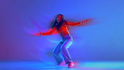 Creative design. Young woman in casual clothes dancing hip hop against gradient blue background in neon light with light wave effect. Concept of dance, art, modern style, youth culture