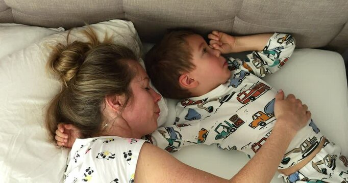 Mother strokes cute little son ribbing eye while lying on double bed at home. Caring woman calms down preschooler son in bedroom. Morning laziness