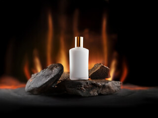 A white perfume bottle with a gold cap rests on top of a pile of dark grey rocks in front of a blurred background of flames.