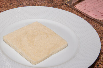 Slice of square bread on a plate next to a blister of sausage before making a sandwich