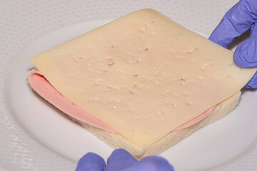Woman hands with nitrile gloves putting a slice of cheese on a slice of mortadella on a sandwich bread