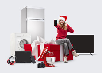 Electronics and appliance store Christmas shopping, smiling woman showing smartphone and thumbs up sitting on big gift box among household appliances, top offers and sales of Christmas shopping online