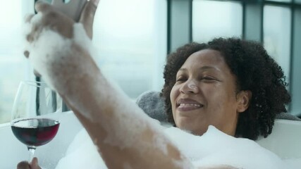 Relaxed Black woman taking selfie on smartphone camera while taking bath with bubbles and red wine in modern bathroom with floor-to-ceiling windows and stunning view on misty city - Powered by Adobe