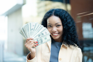 Confident young woman smiling while holding a fan of dollar bills, representing financial success...