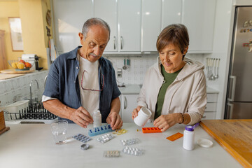 Senior couple managing their medication in warm kitchen. Dressed in casual clothing, organising pillbox as part of their daily routine at home. Horizontal