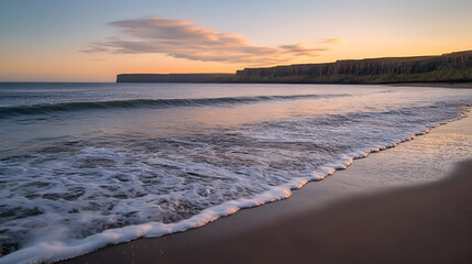 Obraz premium A serene beach scene at sunrise, featuring a calm ocean with gentle waves lapping at the shore and a dramatic cliff face in the distance.