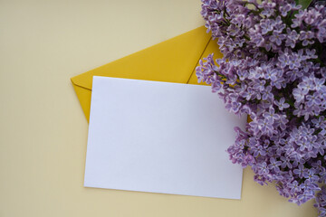 Composition with empty yellow envelope and beautiful spring lilac flowers on beige background. Mockup card invitation greeting card postcard copy space template blank. Branches of lilac blooming