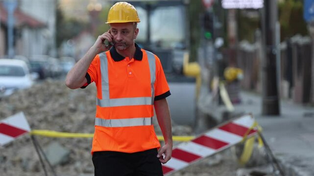 A construction worker wearing a bright orange safety vest and helmet speaks on the phone while monitoring activity in a bustling urban area where construction is underway.