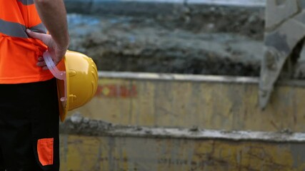 A construction worker in a bright safety vest observes the ongoing work at a construction site. The worker appears to be assessing the project progress while standing near construction materials.