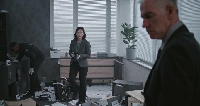 Office robbery case: Crime investigation team at crime scene. Female criminalist takes photos of clues and evidence, then talks to male detective. African American policeman works in the background.