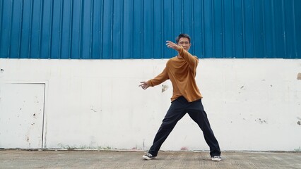 Hispanic man stretch arms and dance street dancing in front of wall. Motion shot of stylish dancer or choreographer in casual outfit practicing dancing in hip hop style. Outdoor sport 2024. Hiphop.