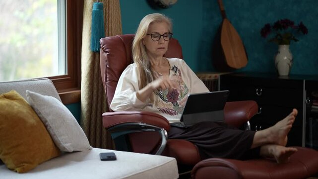 Mature woman in a white blouse and glasses sits in a brown leather recliner, using a tablet computer with her feet up. The room is decorated with a blue wall, a window.