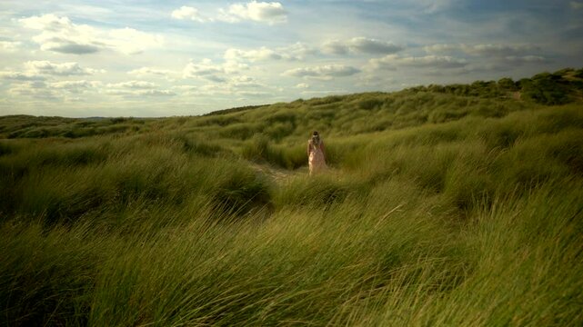 Woman in Pink Dress Walk Through Grass on Beach with Summer Blue Sky Cloud Wind 