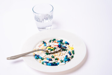 Metal spoon with A lot of Assorted pharmaceutical pills and colorful capsules on a white plate. Glass of water in background.