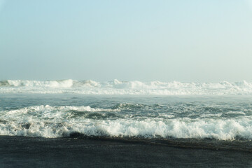 A serene coastal scene with powerful waves crashing onto a dark, sandy shore. The vast expanse of the ocean meets a clear, blue sky, creating a breathtaking panorama