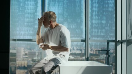 Medium shot of good-looking gray-haired Caucasian adult man dressed in white T-shirt and gray pants sitting on elegant bathtub while applying skincare cream to his face