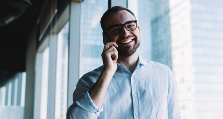 Half length portrait of cheerful male banker in classic eyewear smiling at camera during smartphone communication with corporate partner, happy businessman using mobile device for online conversation