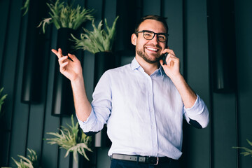 Half length portrait of cheerful formally dressed male entrepreneur in optical eyewear enjoying mobile phone conversation with corporate partner discussing joint stock company, cellular communication