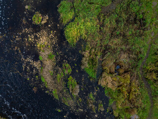 Top-down aerial photo of the Jagala River near Jägala Waterfall in Estonia. On the left the river flows with dark, almost black water speckled with white foam. On the right rugged green landscape.