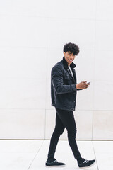 Smiling handsome young curly man in casual outfit browsing mobile while walking on marble passage